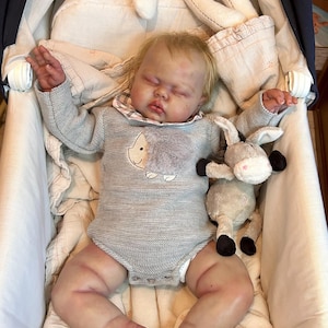 May include: A lifelike baby doll lies in a white bassinet, wearing a gray knit onesie with a sheep applique. A stuffed bunny and donkey are nearby. The doll has blonde hair and is surrounded by soft bedding.