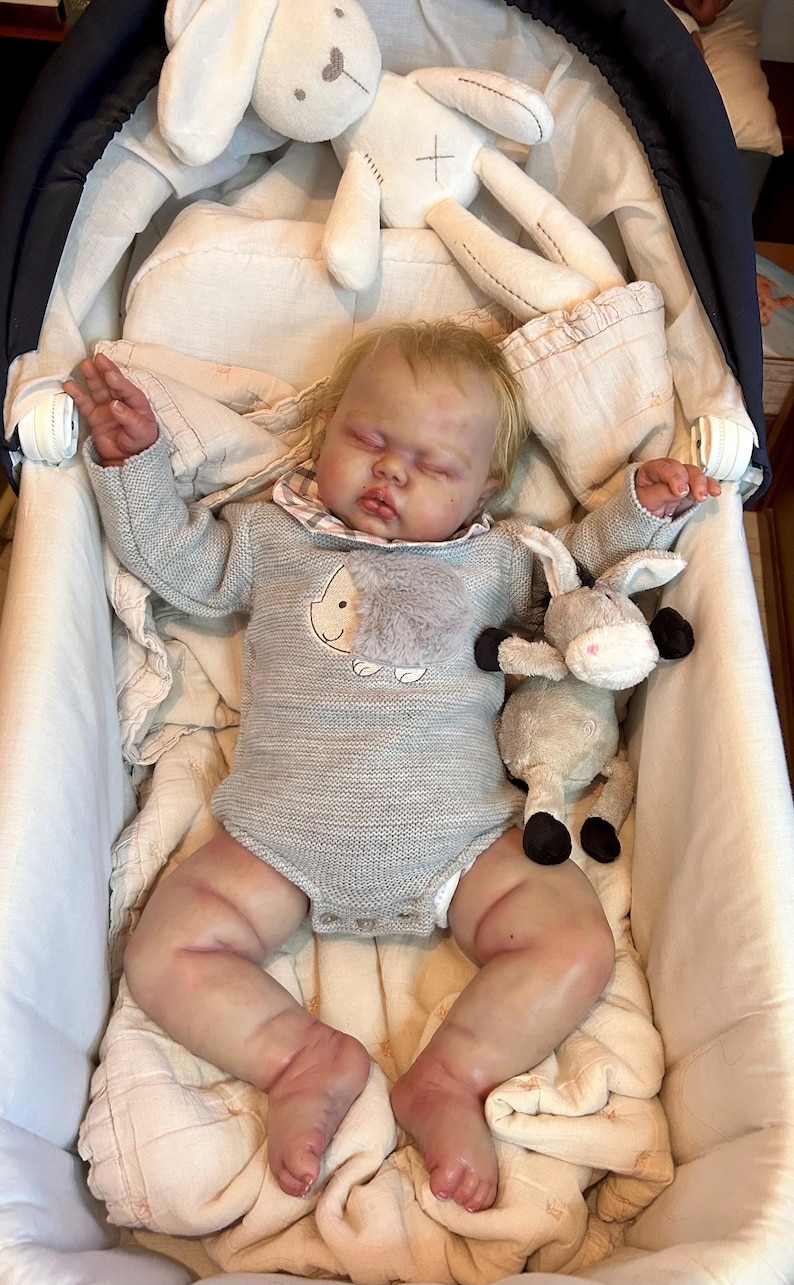 May include: A lifelike baby doll lies in a white bassinet, wearing a gray knit onesie with a sheep applique. A stuffed bunny and donkey are nearby. The doll has blonde hair and is surrounded by soft bedding.