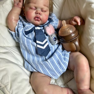 May include: A realistic baby doll wearing a blue and white striped outfit with a navy tie and a plush bunny. The doll has closed eyes and is holding a brown baby bottle. The doll is resting in a white cushioned seat.