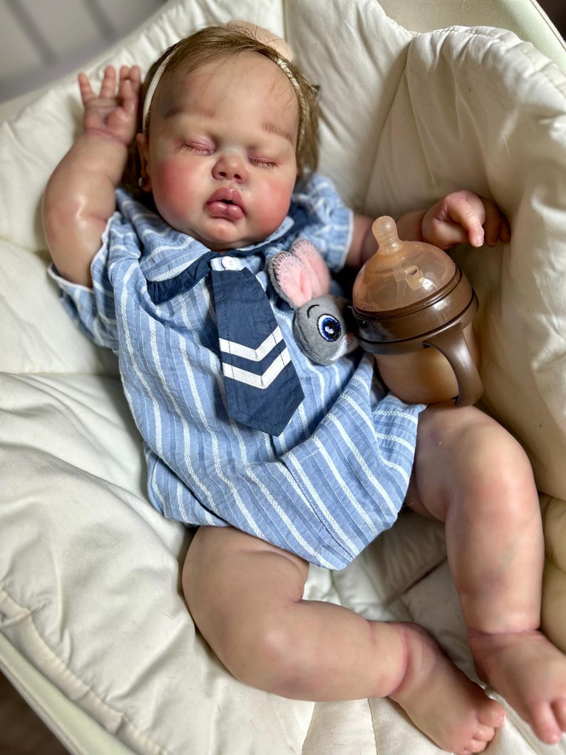 May include: A realistic baby doll wearing a blue and white striped outfit with a navy tie and a plush bunny. The doll has closed eyes and is holding a brown baby bottle. The doll is resting in a white cushioned seat.