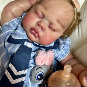 May include: A realistic baby doll with closed eyes, rosy cheeks, and light brown hair. The doll wears a blue and white striped shirt with a navy blue tie and a plush bunny decoration. A baby bottle is also visible.