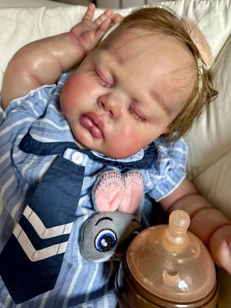 May include: A realistic baby doll with closed eyes, rosy cheeks, and light brown hair. The doll wears a blue and white striped shirt with a navy blue tie and a plush bunny decoration. A baby bottle is also visible.