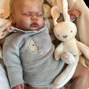 May include: A realistic reborn doll with blonde hair and closed eyes, wearing a grey knit long-sleeve romper with a textured grey patch and a plaid collar. The doll is lying down next to a soft, cream-colored plush rabbit toy with long ears.
