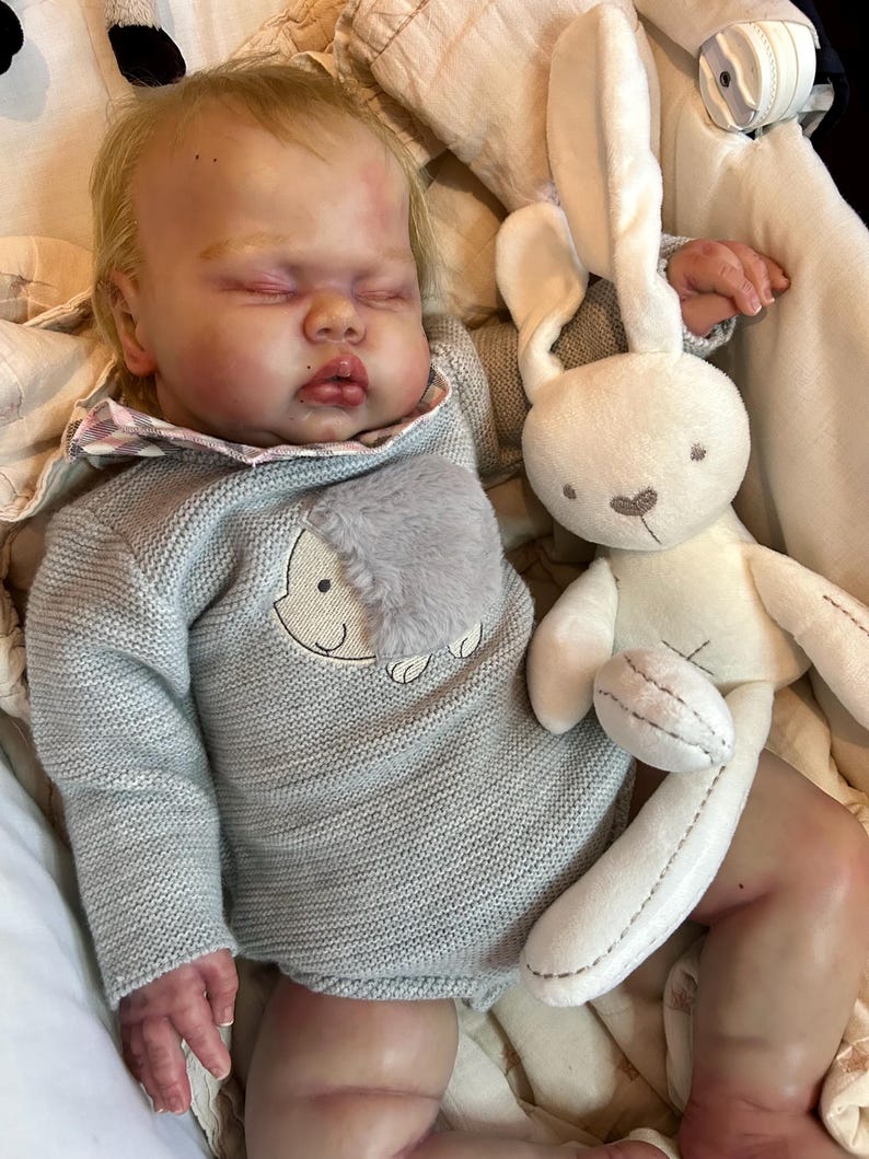 May include: A realistic reborn doll with blonde hair and closed eyes, wearing a grey knit long-sleeve romper with a textured grey patch and a plaid collar. The doll is lying down next to a soft, cream-colored plush rabbit toy with long ears.