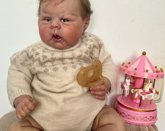 May include: A lifelike baby doll wearing a beige sweater with a patterned yoke, holding a pacifier. A pink carousel toy with white horses is in the background. The doll has realistic skin tones and details.