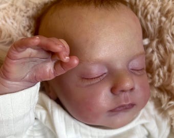 May include: A realistic reborn doll with fair skin, closed eyes, and fine hair, wearing a white long-sleeved shirt with the text "TAY CLUB" printed in grey on the chest. The doll has a hand raised near its face and is resting on a textured surface.