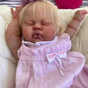 May include: A lifelike baby doll with closed eyes, wearing a light pink dress with white polka dots and a white bow. The doll has blonde hair and a matching pink flower hair accessory. The doll is lying on a white surface.