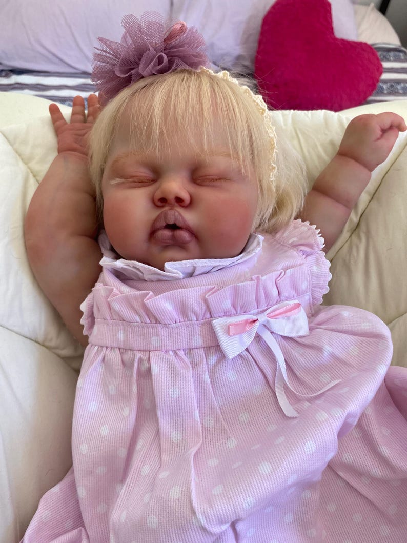 May include: A lifelike baby doll with closed eyes, wearing a light pink dress with white polka dots and a white bow. The doll has blonde hair and a matching pink flower hair accessory. The doll is lying on a white surface.
