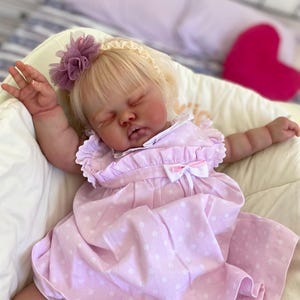 May include: A lifelike baby doll with blonde hair, wearing a pink polka-dot dress and a floral headband. The doll is lying down with its arms outstretched. The dress has a white bow and ruffle details. The doll is in a cream-colored chair.