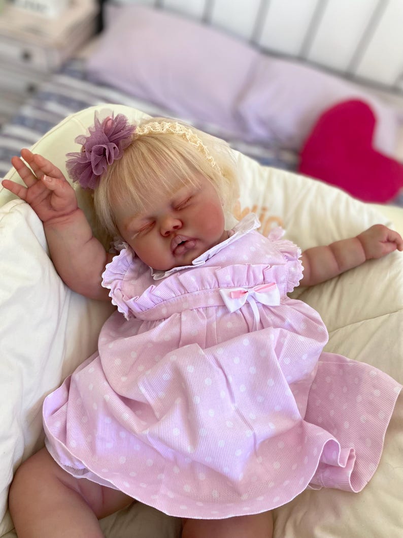 May include: A lifelike baby doll with blonde hair, wearing a pink polka-dot dress and a floral headband. The doll is lying down with its arms outstretched. The dress has a white bow and ruffle details. The doll is in a cream-colored chair.
