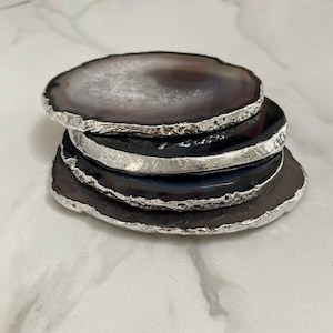 Peut inclure: Ensemble de quatre sous-verres en agate avec une bordure argent&eacute;e. Les sous-verres sont noirs et blancs avec un motif d'agate naturel.