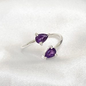 May include: A silver ring with two pear-cut, deep purple gemstones. The gemstones are held in place by prongs, and the ring has an open design. The ring is shown on a white, textured background.