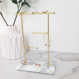 Puede incluir: Un expositor de joyas de metal dorado con base de mármol. El expositor tiene varias barras para colgar collares y pulseras, y una bandeja pequeña para anillos y pendientes.