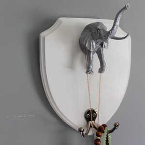May include: A white wooden plaque with a silver elephant head mounted on it. The plaque has two hooks on the bottom, with a necklace and bracelets hanging from them.