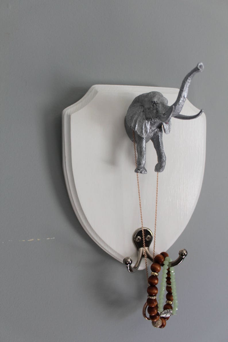 May include: A white wooden plaque with a silver elephant head mounted on it. The plaque has two hooks on the bottom, with a necklace and bracelets hanging from them.