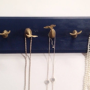 May include: A navy blue wooden wall-mounted jewelry organizer with five gold whale tail hooks. Two necklaces and a pearl strand are hanging from the hooks.