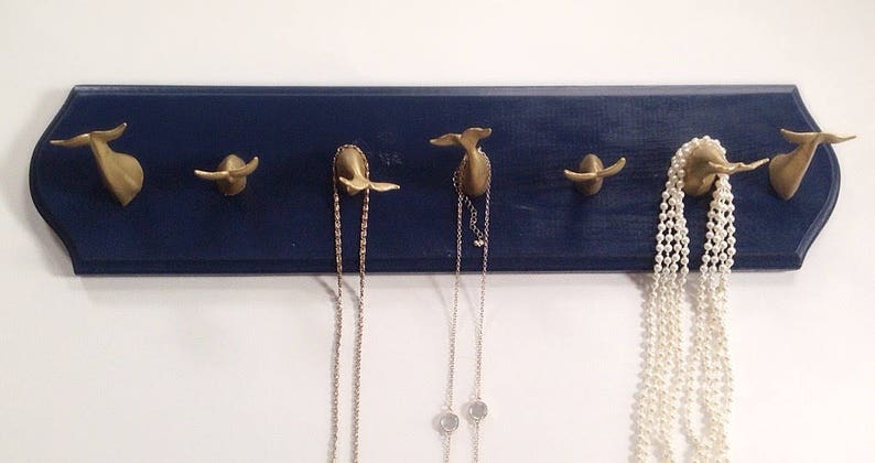 May include: A navy blue wooden wall-mounted jewelry organizer with five gold whale tail hooks. Two necklaces and a pearl strand are hanging from the hooks.