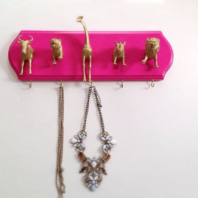 May include: A pink wooden wall-mounted rack with gold animal hooks. The hooks are shaped like a giraffe, a rhino, a lion, and a bull. Two necklaces are hanging from the hooks.