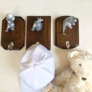 May include: Three wooden wall hooks with silver dinosaur figurines. The hooks are brown and have a distressed finish. The dinosaurs are a triceratops, a stegosaurus, and a tyrannosaurus rex.