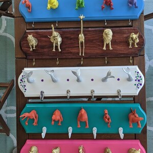 May include: Five wooden coat racks with hooks, each painted a different color. The top rack is blue, the second is brown, the third is white, the fourth is teal, and the bottom is pink. Each rack has a different animal figurine on each hook. The animals include a giraffe, a dinosaur, a rhinoceros, a mermaid, and a horse.