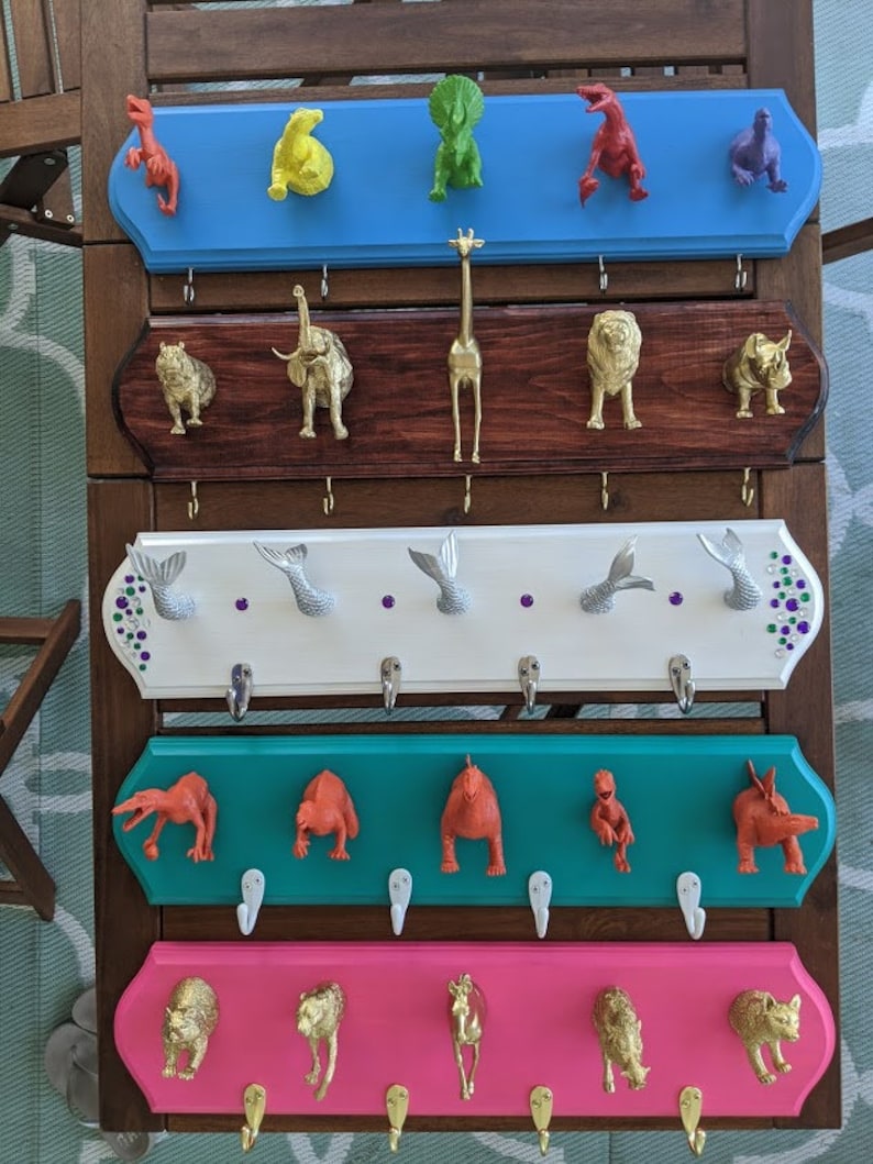 May include: Five wooden coat racks with hooks, each painted a different color. The top rack is blue, the second is brown, the third is white, the fourth is teal, and the bottom is pink. Each rack has a different animal figurine on each hook. The animals include a giraffe, a dinosaur, a rhinoceros, a mermaid, and a horse.