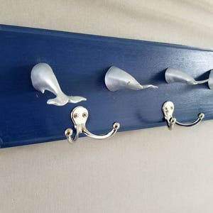 May include: A blue wooden coat rack with five silver whale-shaped hooks. The hooks are attached to a dark blue wooden board with rounded edges.