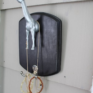 May include: A silver giraffe figurine mounted on a black plaque with a hook for hanging jewelry. A gold chain necklace, a brown beaded bracelet, and a white beaded bracelet are hanging from the hook.
