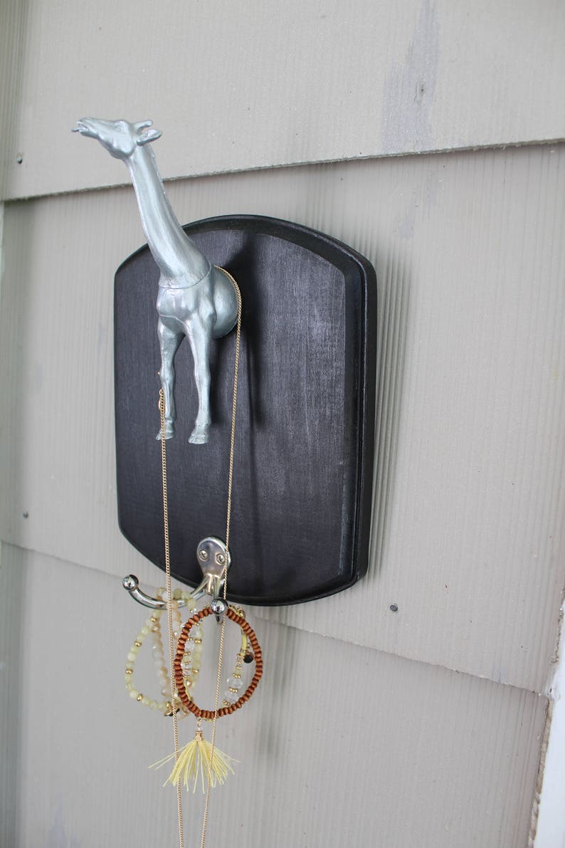May include: A silver giraffe figurine mounted on a black plaque with a hook for hanging jewelry. A gold chain necklace, a brown beaded bracelet, and a white beaded bracelet are hanging from the hook.