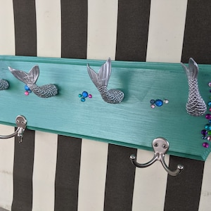May include: A turquoise wooden coat rack with four mermaid tail hooks and silver hooks. The rack is decorated with iridescent gems.