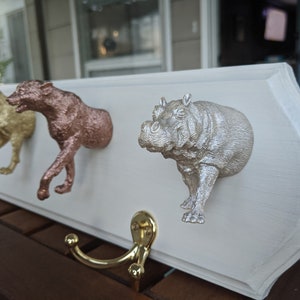 May include: A white wooden coat rack with four animal-shaped hooks. The hooks are a gold dog, a rose gold wolf, a silver hippopotamus, and a silver fox. The rack is mounted on a wall.