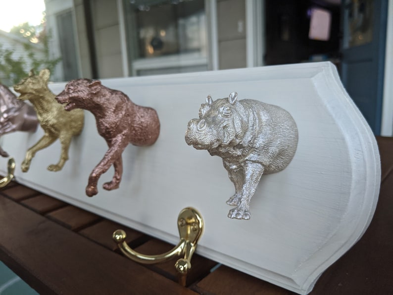 May include: A white wooden coat rack with four animal-shaped hooks. The hooks are a gold dog, a rose gold wolf, a silver hippopotamus, and a silver fox. The rack is mounted on a wall.