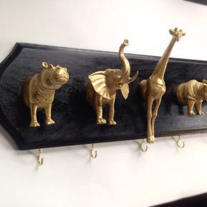 May include: A black wooden coat rack with gold animal hooks. The hooks are shaped like a hippopotamus, elephant, giraffe, and rhinoceros. The rack has five hooks.