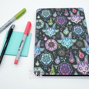 May include: A black notebook with a colorful pattern of goats, pentagrams, and flames. The notebook has a purple border and the text "ICING ON THE STITCH" is printed on the bottom.