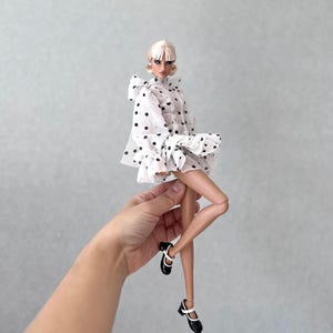 Op de afbeelding: Een modepop in een witte jurk met zwarte stippen. De pop heeft blond haar en draagt zwart-witte schoenen. De jurk heeft een geplooid zoom en een grote strik. De pop wordt door een hand tegen een grijze achtergrond gehouden.