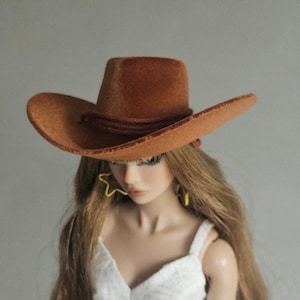 Puede incluir: Un sombrero de vaquero marrón con ala ancha y una banda marrón. El sombrero está sobre una muñeca con cabello largo y castaño claro y un vestido blanco. La muñeca lleva pendientes amarillos en forma de estrella.