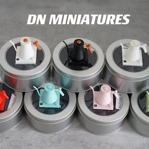May include: Seven miniature ceramic teapots in different colors, displayed in round metal tins. The tins are stacked in a pyramid shape. The text "DN MINIATURES" is printed in white on a black background above the tins.