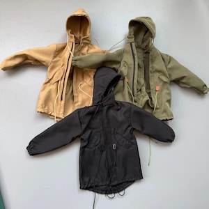 May include: Three miniature hooded jackets in tan, olive green, and black. Each jacket has a front zipper, pockets, and drawstrings. The jackets are designed for a doll or action figure.