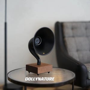 May include: A black vintage-style gramophone speaker on a wooden base sits on a round glass table with a black metal frame. The speaker is on a table in a room with a black armchair and a black floor lamp.