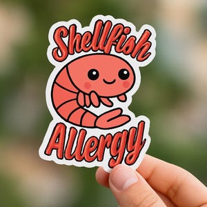 May include: A sticker with a cartoon shrimp design and the words "Shellfish Allergy". The shrimp is red with black details and a smiling face. The text and shrimp are outlined in white. The sticker is held by a hand.