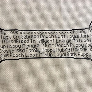 May include: A black and white cross-stitch design of a dog bone with the words "Puppy, Mutt, Woof, Eager, Mongrel, Family, Love, Happy, Mixed Breed, Fur, Intelligent, Energetic, Excitable, Crossbreed, Pooch, Coat, Loyal, Bark, Hybrid, Dog, Pup, Mixup, Fun, Dog Coat, Love, Mutt Dog" written around the bone.