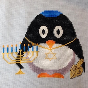 May include: A black and white penguin wearing a blue yarmulke and a gold necklace with a Star of David pendant. The penguin is holding a menorah with lit candles and a dreidel.