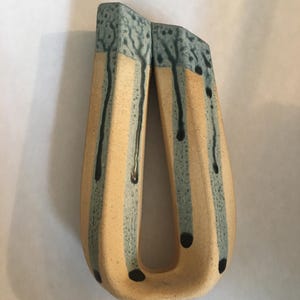 May include: A unique ceramic vase with a U-shaped design. The vase features vertical stripes in beige and light blue, with dark blue drips and black dot accents. The top edges are textured light blue. A decorative piece for the home.