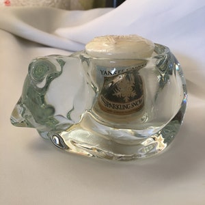 May include: A clear glass candle holder shaped like a bear, holding a small white candle with a gold label that reads "Yankee Candle Sparkling Snow with authentic essential oils".
