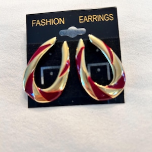 May include: Gold-tone earrings with a teardrop shape and red enamel accents. The earrings are displayed on a black card that reads "FASHION EARRINGS". The earrings have a glossy finish and a modern design.