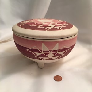 May include: A ceramic pot with a lid, decorated with a geometric pattern in shades of pink and white. The pot has three legs and is sitting on a white surface.