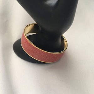 May include: A coral-colored bangle bracelet with a gold-toned rim and a white floral pattern. The bracelet is on a black display stand.
