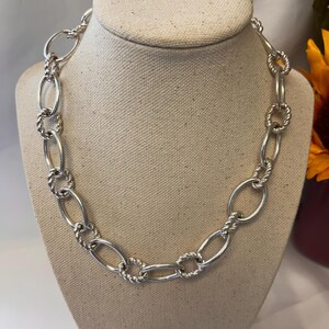 May include: A silver-toned necklace with a chunky chain design. The necklace features oval links connected by twisted and circular links. The jewelry is displayed on a beige mannequin head.