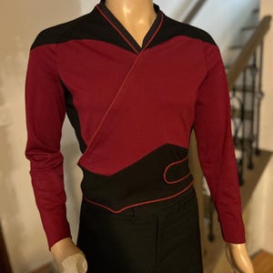 Op de afbeelding: Een lang mouw shirt met een bordeauxrode en zwarte kleurstelling. Het shirt heeft een v-hals en een rode rand rond de randen. Het shirt is op een mannequin.