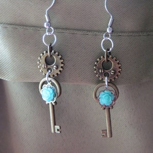 Può includere: Un paio di orecchini in stile steampunk. Ogni orecchino presenta un gancio argentato, un ingranaggio color bronzo, un ciondolo a forma di chiave e un fiore azzurro. Gli orecchini sono sospesi su uno sfondo neutro.