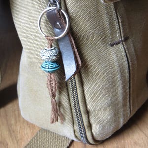 Può includere: Primo piano di una borsa in tela beige con un portachiavi decorativo. Il portachiavi presenta due perline decorative, una bianca con un motivo floreale e una turchese con un motivo geometrico. La borsa ha un cinturino in pelle marrone.